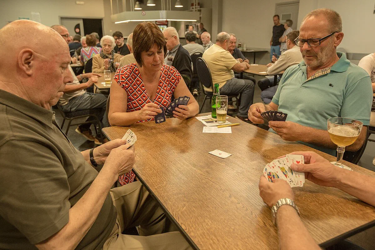 Quatre joueurs autour d'une table en bois pendant un tournoi de belote, cartes en main, une carte posée au centre et une feuille de marque à côté des verres