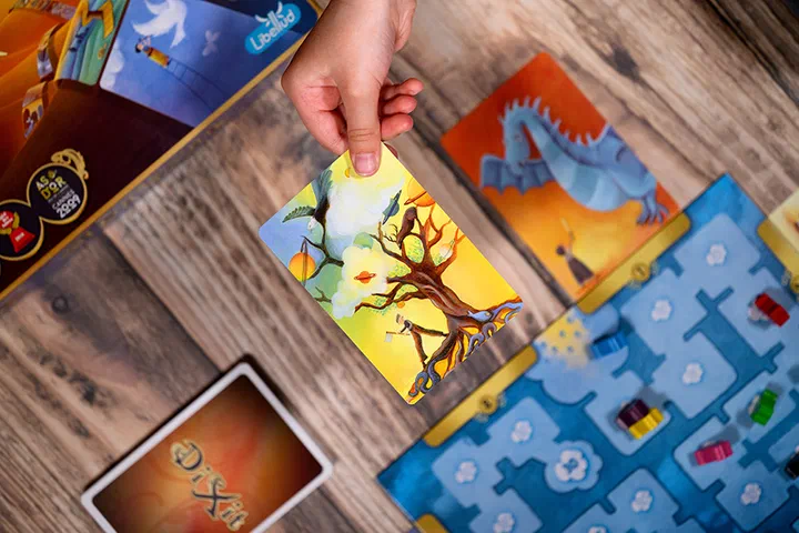 Partie de Dixit : une main tient une carte illustrée d'un arbre aux planètes au-dessus du plateau bleu où avancent les lapins en bois